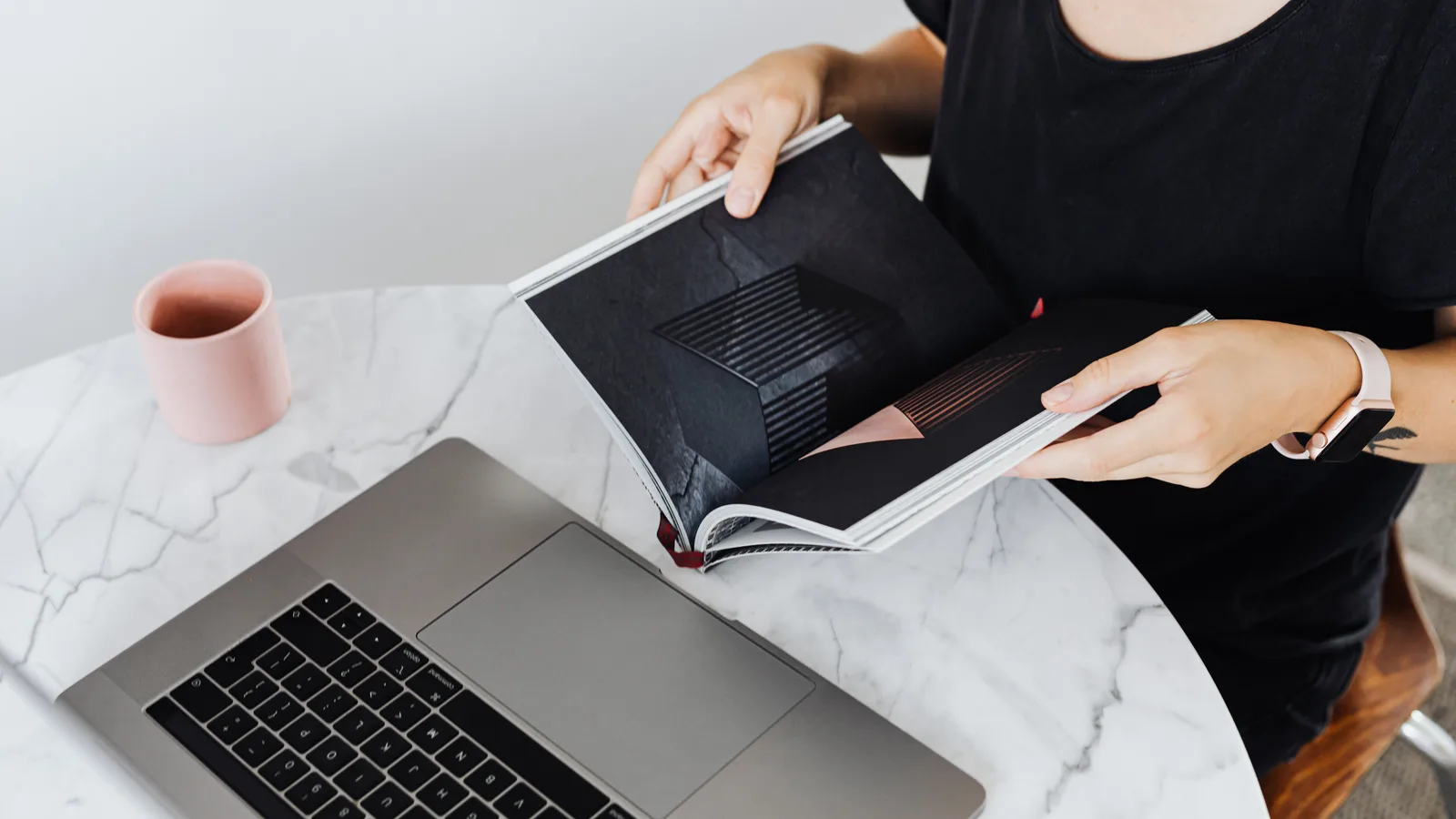 A woman reads a book at a marble table with a laptop and coffee cup in a modern workspace.