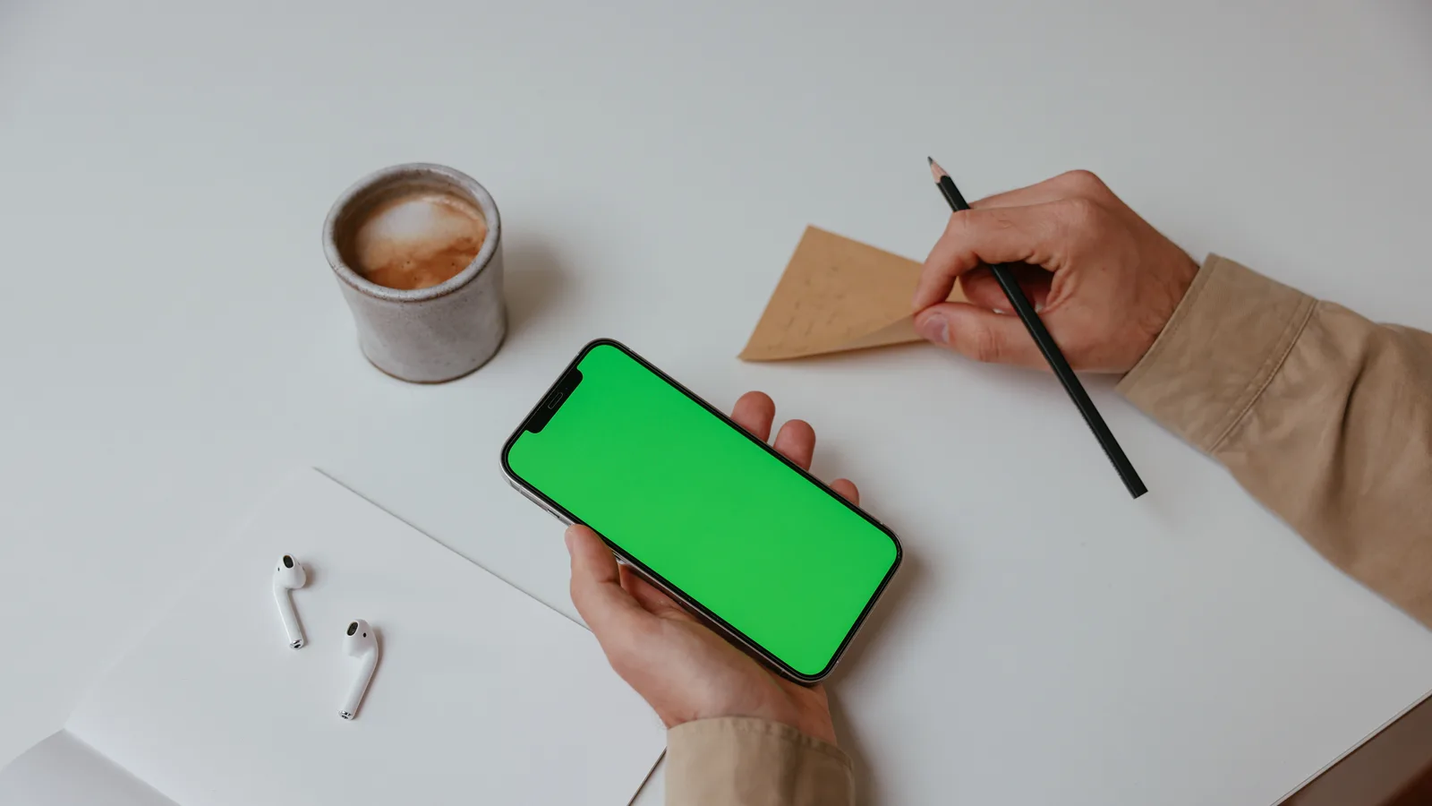 A minimalist workspace featuring a smartphone with a green screen, coffee, and stationery on a white desk.